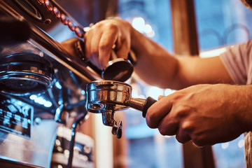 Close-up photo of a barista holding a portafilter with a black ground coffee and working on a coffee machine in a cafe shop or restaurant.