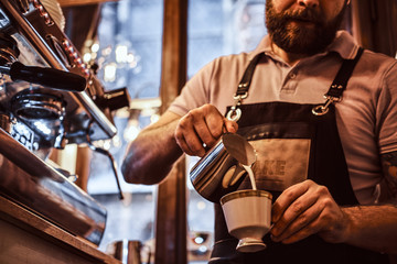 Barista in apron making a cappuccino, pouring milk in a cup in a restaurant or coffee shop