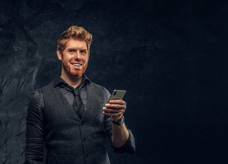 Cheerful redhead man in formal wear holding a smartphone in studio against a dark textured wall