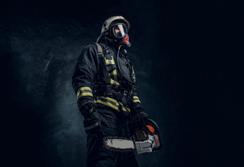 Firefighter in safety helmet and oxygen mask holding a chainsaw. Studio photo against a dark textured wall