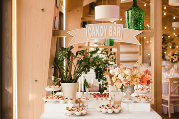 Candy bar with tasty desserts and appetizers at wedding reception. Decorated with flower compositions and candles. Sweet food, dessert, buffet, catering table, restaurant.