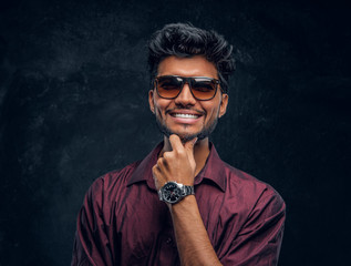 Vogue, fashion, style. Cheerful young Indian guy wearing a stylish shirt and sunglasses posing with hand on chin. Studio photo against a dark textured wall