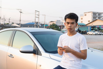 A boy iin white casual is chatting on his phone and leaning on the silver car hood.