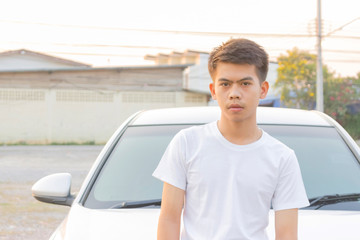 A boy iin white casual is standing in front of the silver car in the evening light