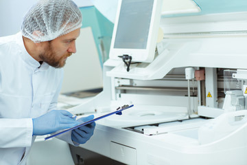 Young doctor in blue gloves working with new equipment. Laboratory assistant with red beard doing analysis. Attractive medical worker in lab coat standing in laboratory and writing results.