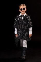 Little girl in a black glasses, dark poncho, black boots and a clutch in her hand