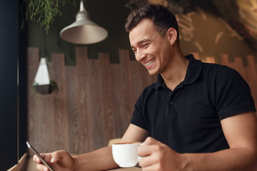 Cheerful guy with coffee browsing smartphone in cafe