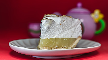 A slice of lemon merigue pie with a toy tea set on a red background.