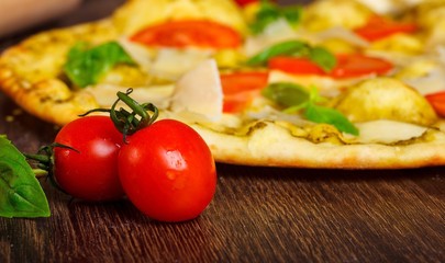 Close up vegetarian Italian pizza with melted mozzarella, parmesan cheese, red tomatoes and fresh green basil leaves on a brown table decorated by mozzarella, hot chili pepper, cherry and battledore