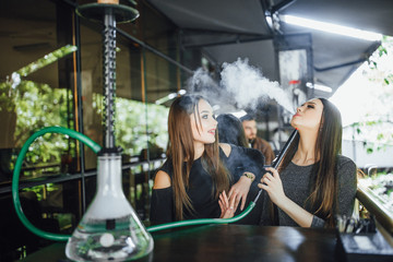 Two beautiful brunette friends are sitting on the summer terrace of a modern restaurant, chatting and smoking hookah, covered with blankets.