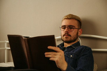 Man reading  book. Young bearded male sitting on couch and is studying literature intently while leaning on wall