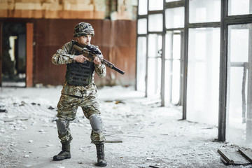 Patrols the territory! The young soldier in the military stands at the window of the collapsed building! On the head is a protective helmet! There is a big gun in his hands!