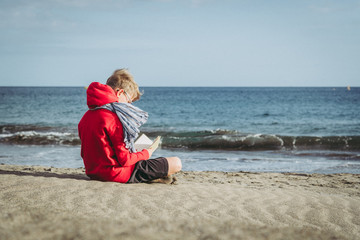 Fototapeta premium Cute boy in a red hoodie reading a book at the beach in a cloudy day, enjoying holiday. Young male sitting on the sand and relaxing with a blast. Kid enjoying a trip in a new exotic place near the sea