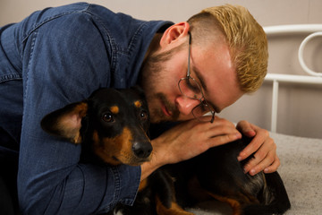 Man hugging dachshund puppy. Calm guy lying with dog on sofa in room.