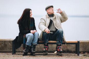 The original pensioner and his daughter are sitting on a bench, resting from roller skating. An elderly man drinks water. Active lifestyle.