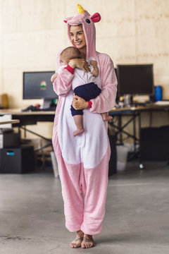 Young mother wearing unicorn onesie, standing in office, holding her son in her arms