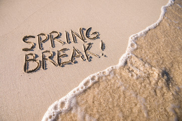 Fototapeta premium Excited Spring Break message handwritten on the smooth sand of an empty beach with an oncoming wave