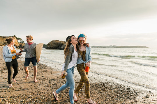 Happy friends with surfboard and drinks walking on stony beach