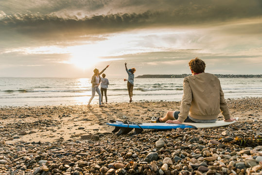 Young man on surfboard watching friends making soap bubbles on the  beach at sunset