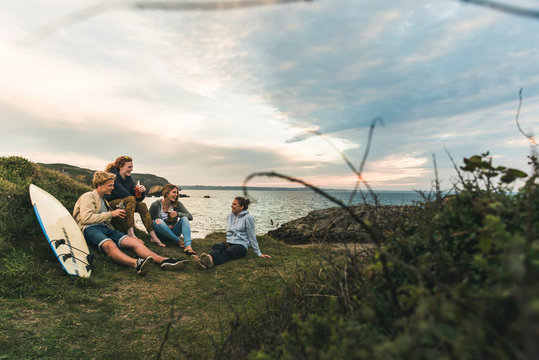 Happy friends with surfboard and drinks socializing at the coast at sunset