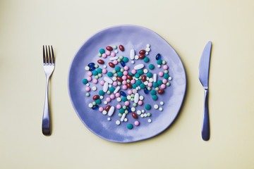 colorful pills, capsules and supplements on ceramic plate, fork and knife on yellow background, top...