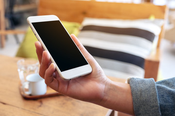Mockup image of a hand holding white mobile phone with blank screen with coffee cup on wooden table in cafe