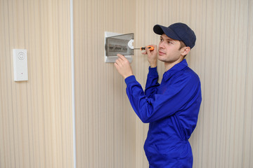 lateral view of a young eletrician in blue overall disassembling a electrical panel with fuses in a house.