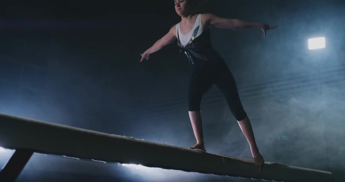The girl is a professional athlete performs gymnastic acrobatic trick on a beam in backlight and slow motion in sports gymnastic clothing. Smoke and blue. Jump and spin on the balance beam