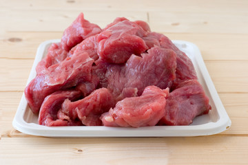 Raw fresh beef meat pieces on a white tray on a a beige plank wooden table. Close up