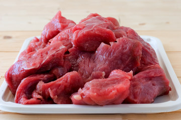 Raw fresh beef meat pieces on a white tray on a a beige plank wooden table. Close up