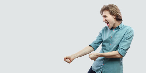 Profile side view portrait of screaming handsome long haired blonde young man in blue shirt standing with boxing gesture or pulling visualization. indoor studio shot, isolated on light grey background