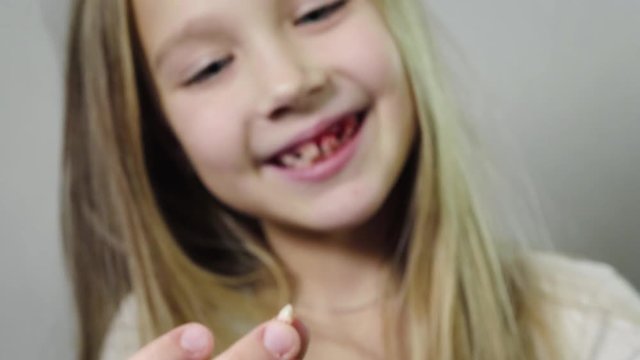 Close - up of a cute smiling girl, a child showing a torn milk Fang in her hand and a bleeding gums, jaw in a room with a white background. Tooth loss by age concept. 4K resolution