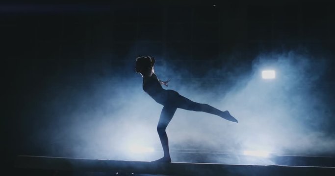 The girl is a professional athlete performs gymnastic acrobatic trick on a beam in backlight and slow motion in sports gymnastic clothing. Smoke and blue. Jump and spin on the balance beam