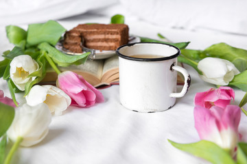 Fototapeta na wymiar mug, tea, white and pink tulips, a piece of cake, a book on a white sheet