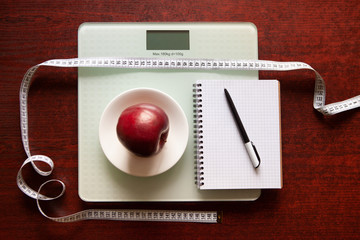 The concept of healthy nutrition, fitness and weight loss. Weights, measuring tape, apple, open notebook and pencil on the table.