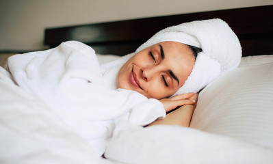 Fototapeta na wymiar Middle day nap. Cute young girl is having a relaxing nap after shower in cozy double bed with bright white bedclothes. Her skin is tanned, eyebrows are nicely shaped and her smile is pleasant.
