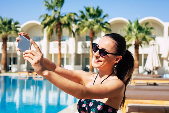 Follow me! Hilariously beautiful girl is taking a selfie next to the swimming pool ,greenish palms,white building and wooden lounge chaises.