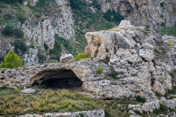 Fototapeta na wymiar Distant soft colors landscape view of the arched entrance of cave under the ancient town of Matera, the Sassi di Matera, Basilicata, Southern Italy, cloudy summer afternoon just before sunset