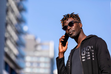 Front view of black man with sunglasses standing against cityscape on the street while using a mobile phone in sunny day.