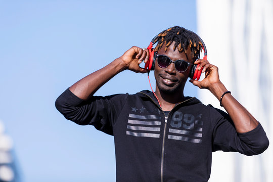 Front view of a black man wearing casual clothes and sunglasses standing in the street while using headphones to listen music in sunny day