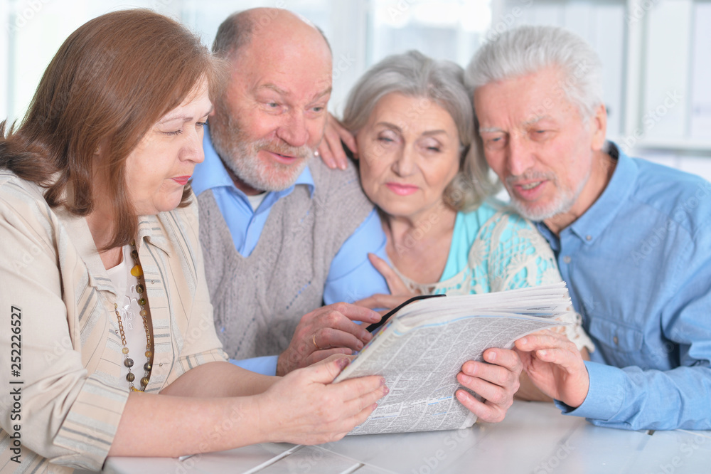 Poster Portrait of two senior couples sitting at table and reading newspaper