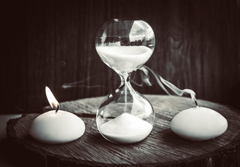 Time running out, life and death concept. One candle burning with flame as living other is blown away as dead and hourglass measures lifetime. 