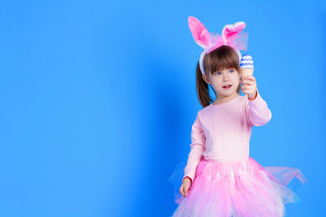 Obraz na płótnie Canvas Cute pink young girl child daughter wears pink dress like rabbit playing in easter holiday game with rabbit ears and ester eggs on isolated blue background. Spring is coming and Easter holiday concept