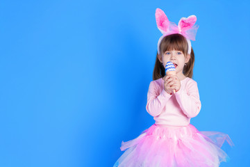 Obraz na płótnie Canvas Cute pink young girl child daughter wears pink dress like rabbit playing in easter holiday game with rabbit ears and ester eggs on isolated blue background. Spring is coming and Easter holiday concept