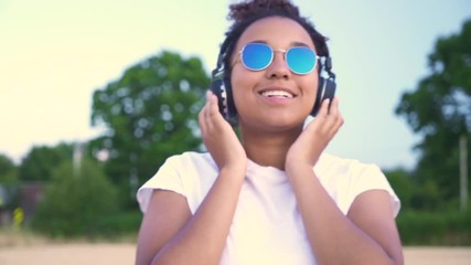 Slow motion follow shot video of beautiful mixed race African American girl teenager young woman in white T-shirt and blue sunglasses walking listening to music on wireless headphones 