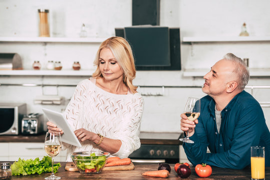 handsome man holding glass with wine and looking at attractive blonde wife holding digital tablet in kitchen