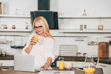 attractive woman in eye glasses drinking orange juice near laptop