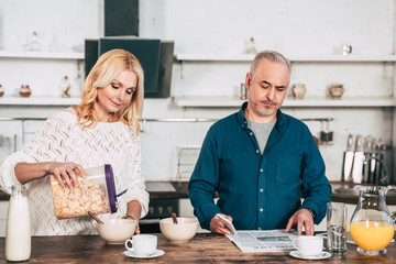 handsome man reading newspaper near wife with cornflakes standing in kitchen