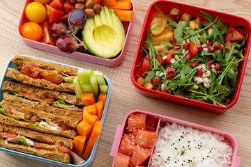Creative layout with healthy lunch dishes variety in bento boxes on wooden table. Sandwich, slad with grains and pomegranate seeds, salmon with rice and fruit snacks. Office or school lunch concept