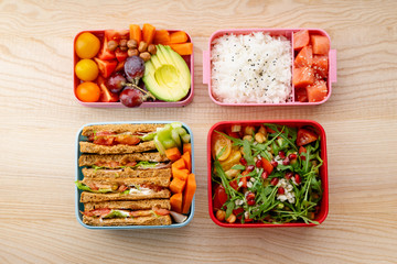Creative layout with healthy lunch dishes variety in bento boxes on wooden table. Sandwich, slad with grains and pomegranate seeds, salmon with rice and fruit snacks. Office or school lunch concept